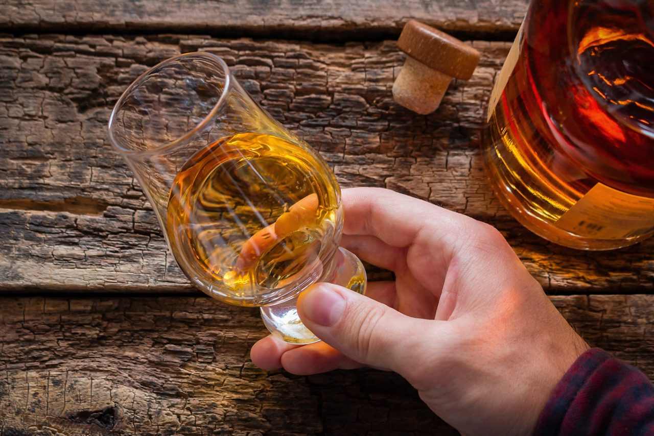 Close-up of a hand holding a glass of amber rum next to a cork and bottle on rustic wooden table in Martinique