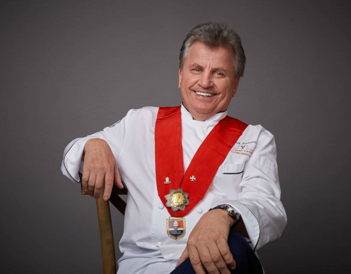 Chef in white uniform with red sash smiling while leaning on wooden handle. 