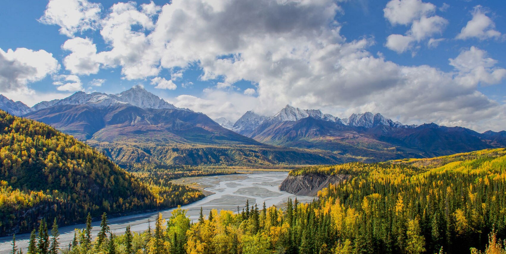 An aerial or elevated view of Matanuska Glacier appearing as a wide white ice field in a valley surrounded by dark forested slopes and tall mountain peaks under cloudy skies.