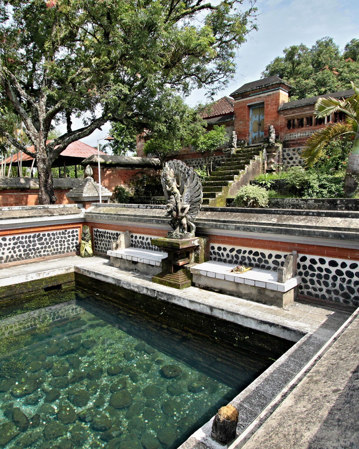 Stone-lined temple pool in Mataram with clear water, a central guardian statue and surrounding brick walls under leafy trees.