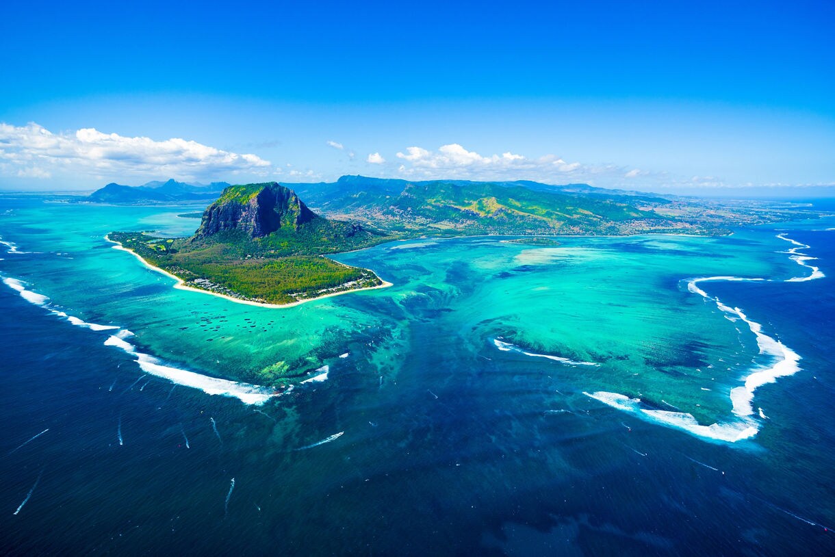 Aerial view of Le Morne Brabant in Mauritius surrounded by bright turquoise lagoon waters, shallow reefs and deep ocean blue under a clear sunny sky.