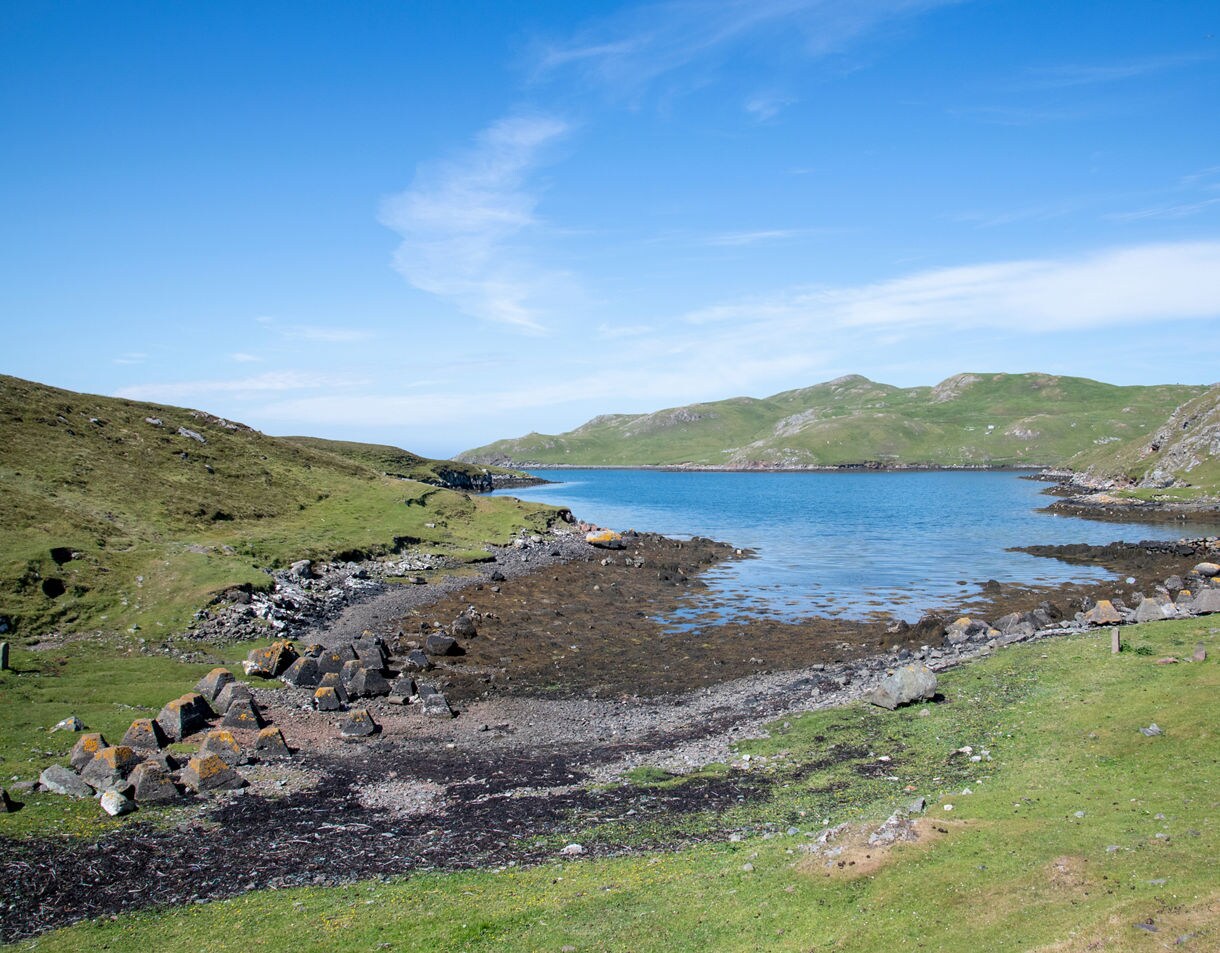 Rocky shoreline and grassy hills at Mavis Grind in Shetland, with a narrow strip of land dividing blue sea waters.