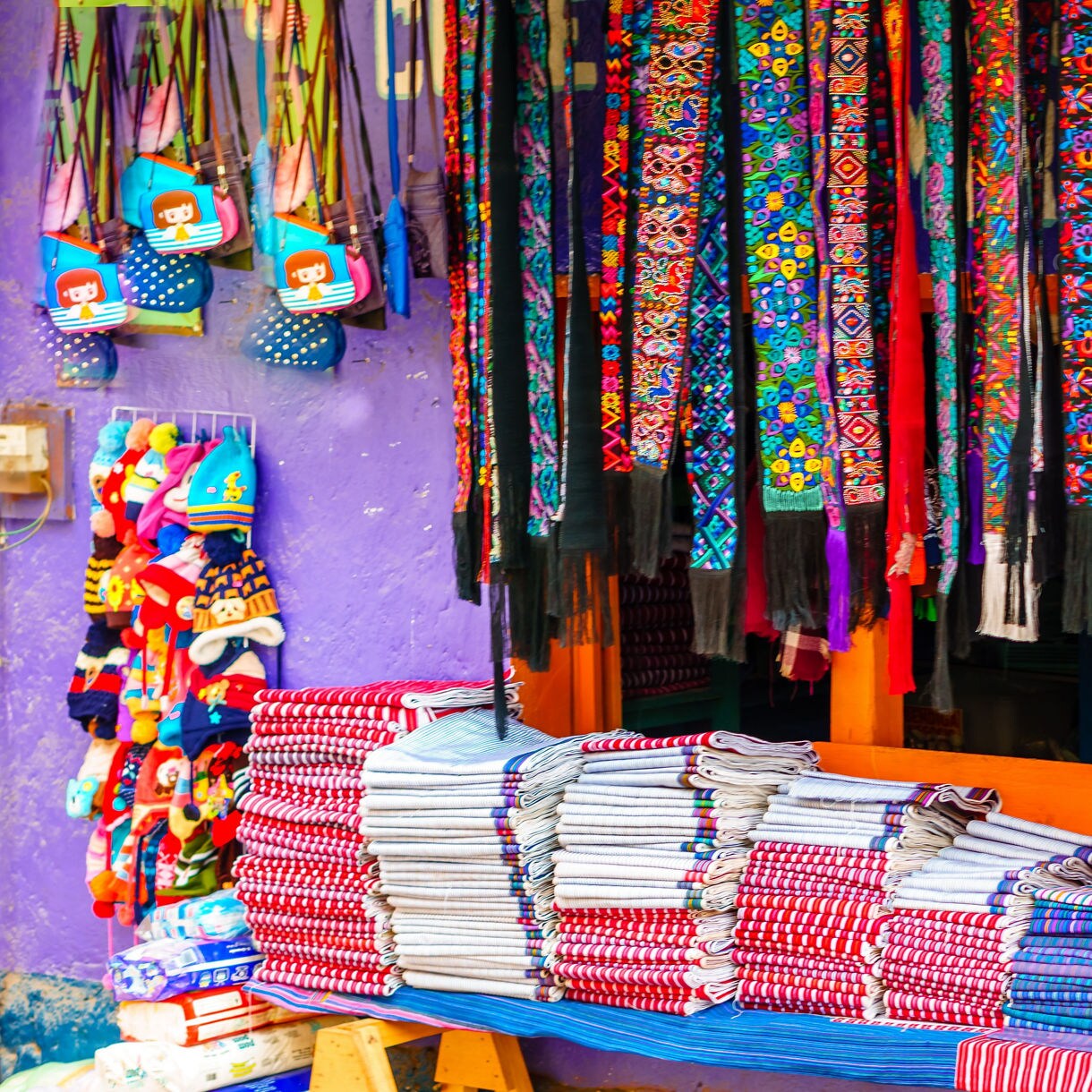 Colorful handmade fabrics, scarves and hats displayed outside a market stall with a purple wall in Guatemala.