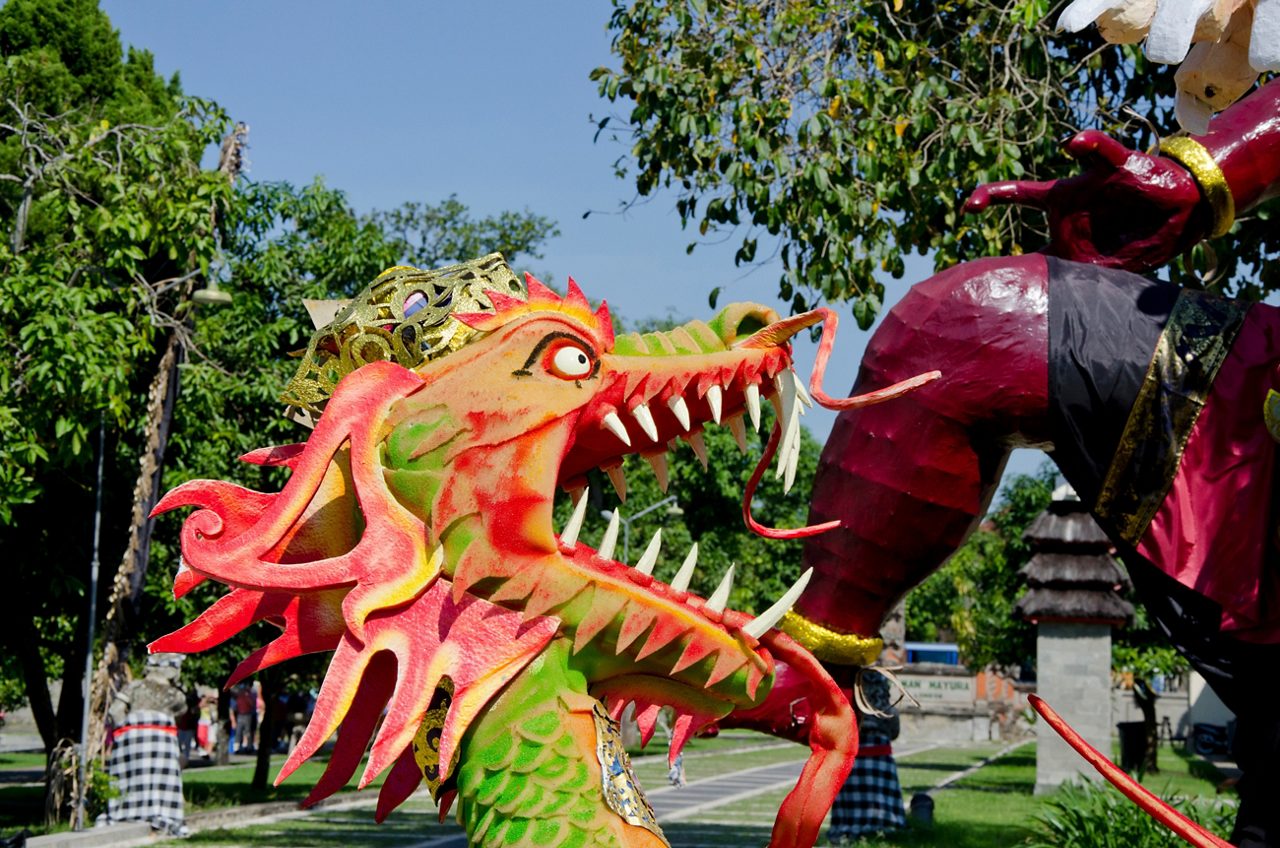 Vibrant close-up of a colorful dragon statue with sharp white teeth and flowing red and yellow details, displayed outdoors near other large ceremonial figures at Mayura Water Palace in Lombok.