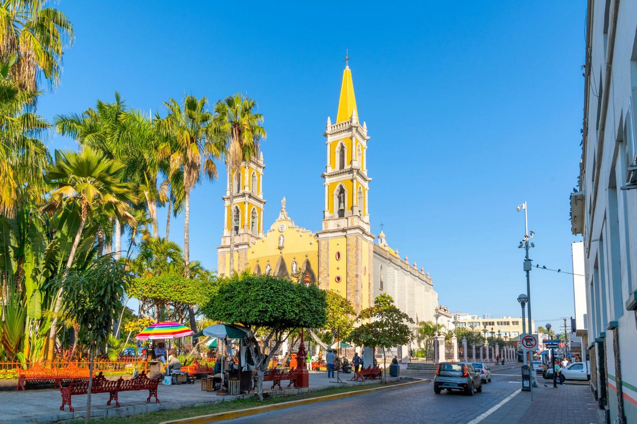 A picturesque view of the Cathedral Basilica of the Immaculate Conception in Mazatlán, Mexico, with its bright yellow towers rising above palm trees in a vibrant town square, with people enjoying the shaded benches and vendors selling goods under colorful umbrellas.