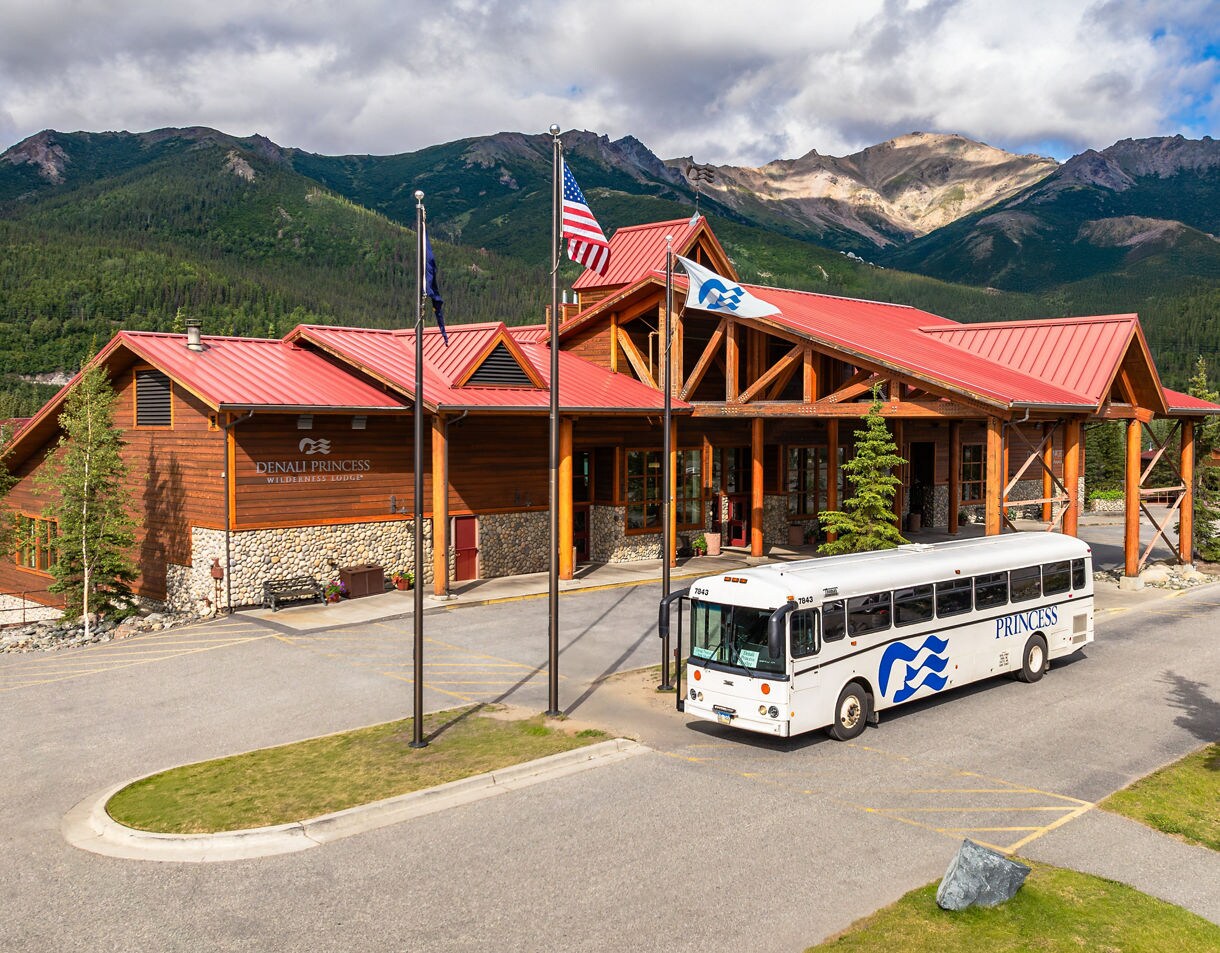 Denali Princess Wilderness Lodge main building exterior with log cabin construction, red metal roof, American and Alaska flags, white Princess tour bus parked in circular driveway, and Alaska Range mountains in background.
