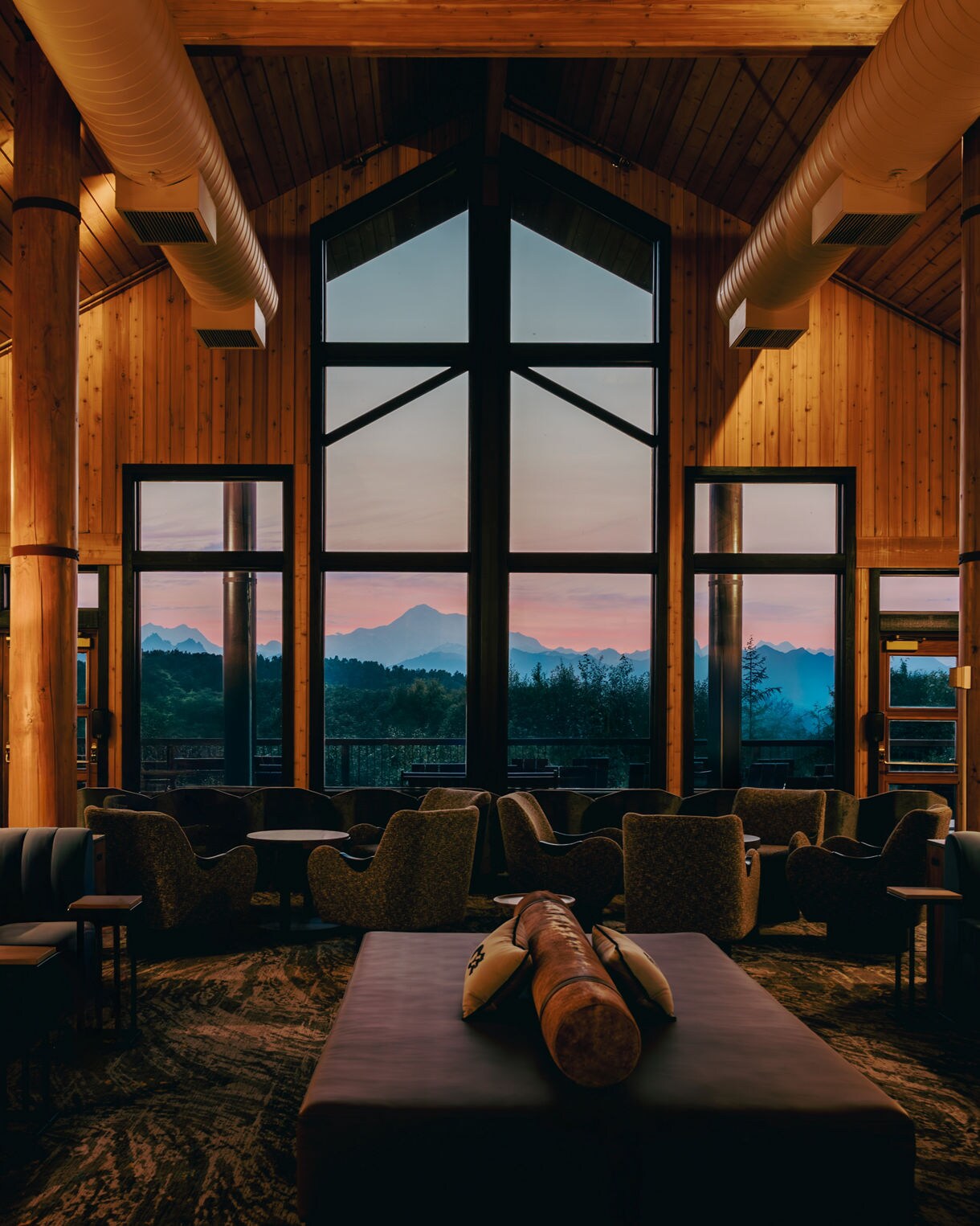 Lodge great room interior with dramatic vaulted wood ceiling and towering windows showcasing layered mountain silhouettes at sunset, with comfortable seating area and leather ottoman in foreground.
