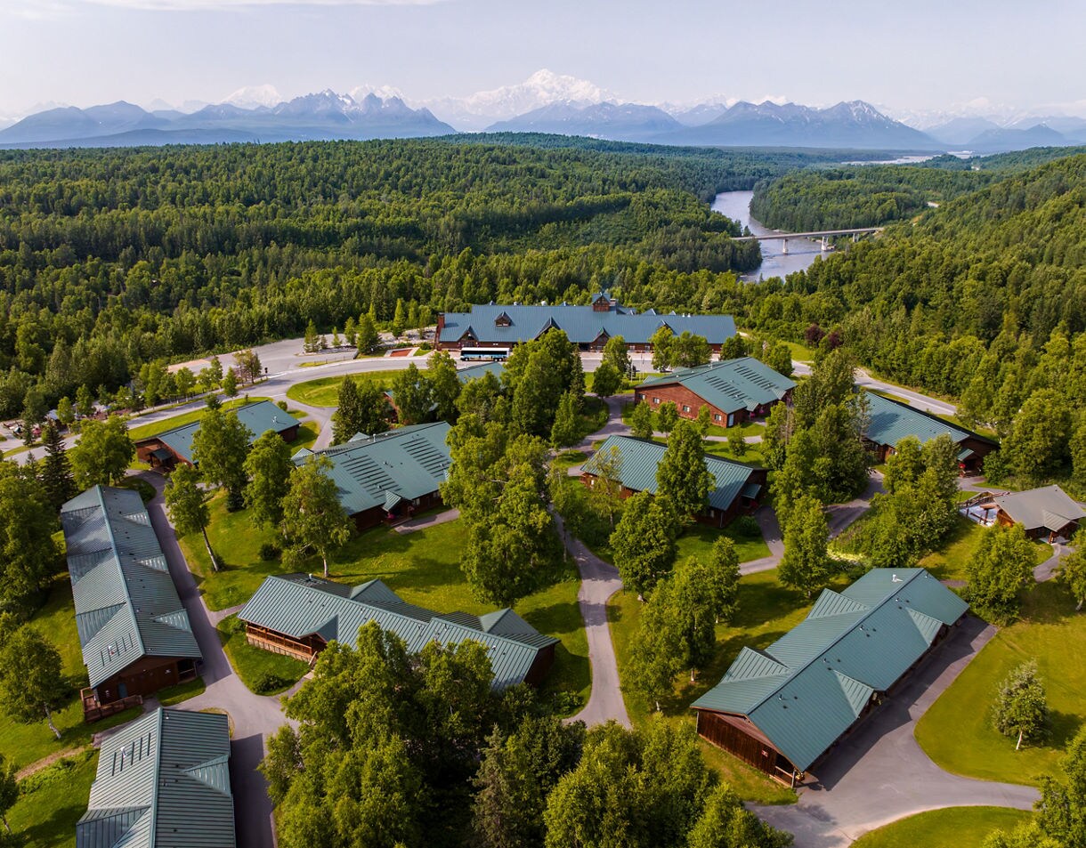 Aerial view of McKinley Princess Wilderness Lodge showing multiple buildings with green metal roofs surrounded by dense boreal forest, with a winding river and snow-capped mountain range visible in the distance.