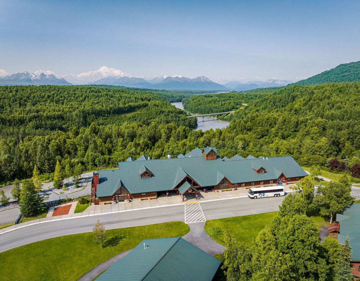 Aerial view of McKinley Princess Wilderness Lodge showing multiple buildings with green metal roofs surrounded by dense boreal forest, with a winding river and snow-capped mountain range visible in the distance.