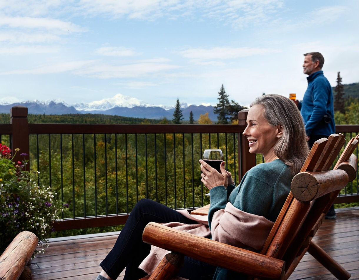 Woman relaxing in rustic wooden Adirondack chair on lodge deck holding wine glass, with man standing in background and forested valley and distant mountains stretching to horizon.