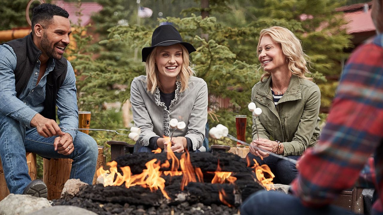 Three people laughing and roasting marshmallows around outdoor fire pit at Denali Princess Wilderness Lodge with evergreen trees in background.
