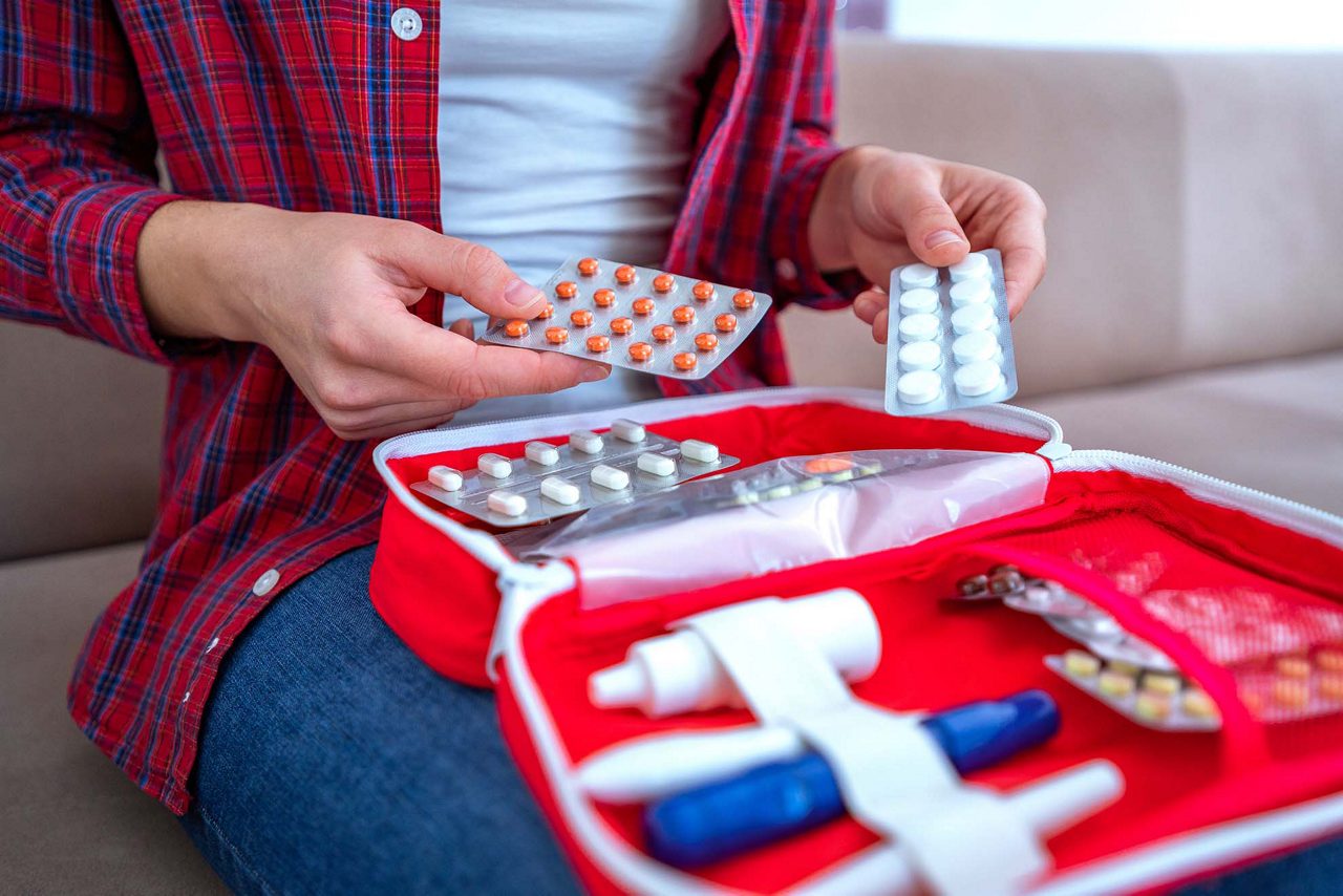 A person organizing blister packs of medication into an open red first-aid travel kit on their lap.