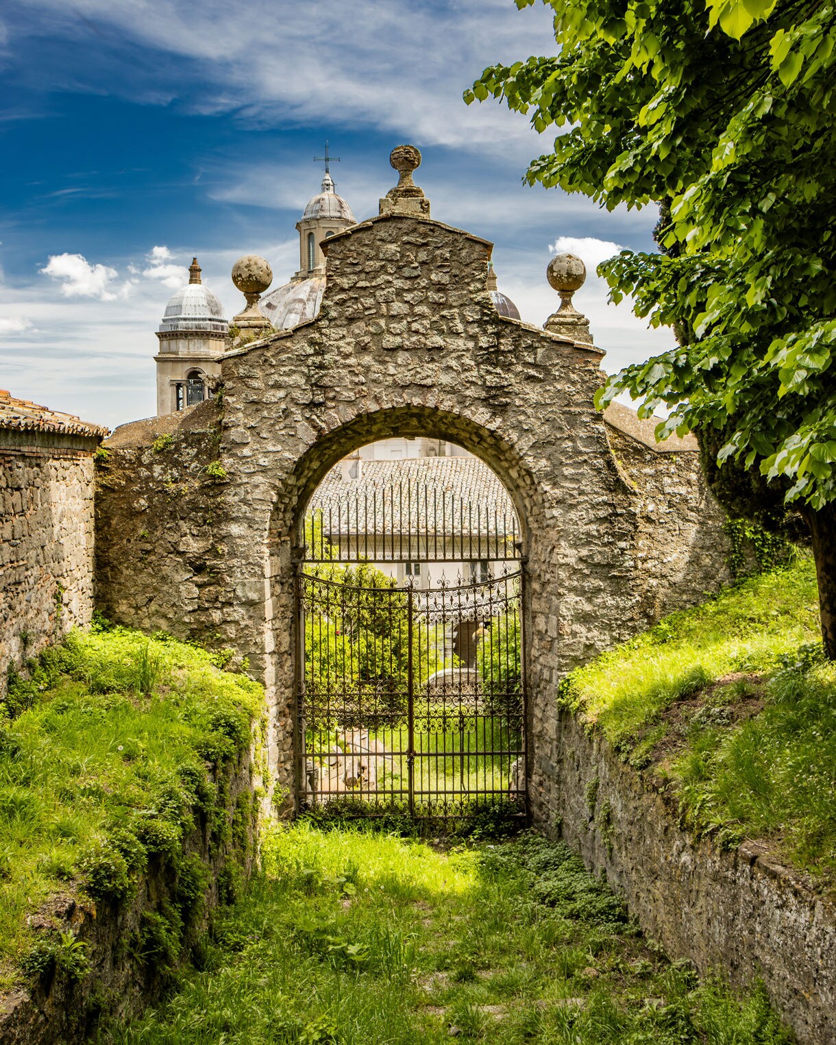 A weathered stone archway with an iron gate stands along a grassy path, framed by leafy trees, with domed church rooftops visible in the distance.