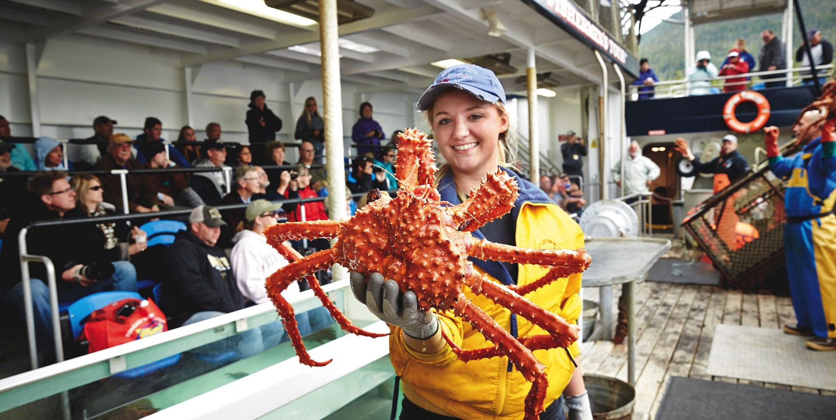 A person in a yellow jacket holds a large red crab on the deck of a tour boat. The scene takes place in front of an audience seated in bleachers, with water tanks and fishing equipment visible. The setting suggests a marine wildlife demonstration or fishing excursion, with a lively and educational atmosphere. The background includes life rings and a partially covered deck area.