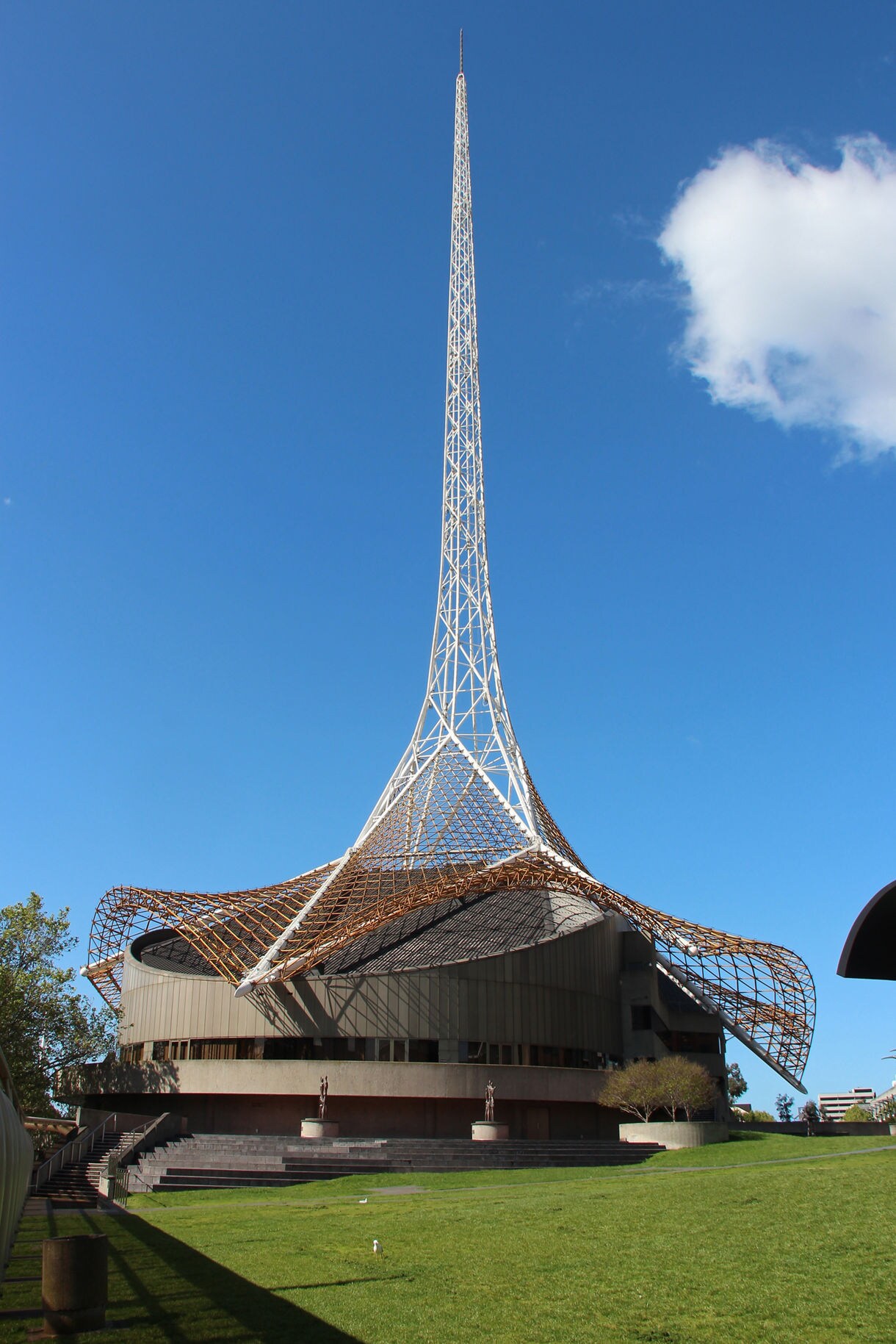 The Melbourne Arts Centre building with its tall white spire and sculptural roof design against a clear blue sky.