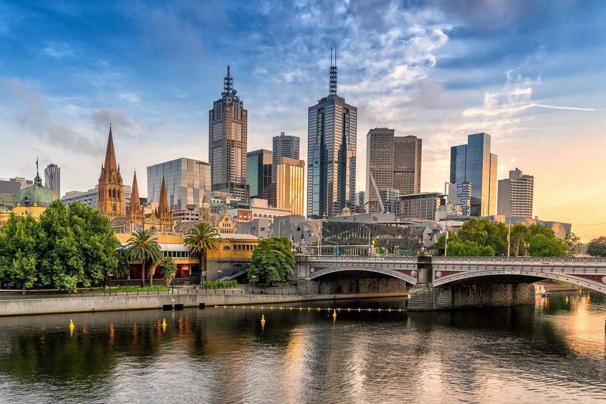 View of Melbourne’s skyline with modern skyscrapers, historic buildings, and the Yarra River reflecting the golden light of sunrise.