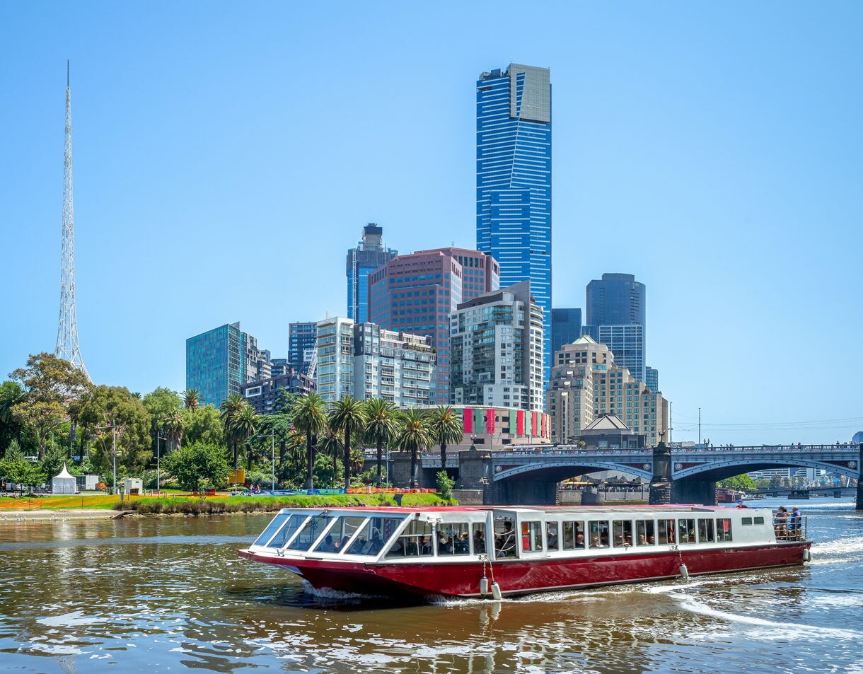 Sightseeing boat cruising on the Yarra River with Melbourne’s modern skyline, palm trees, and bridges under a clear blue sky.