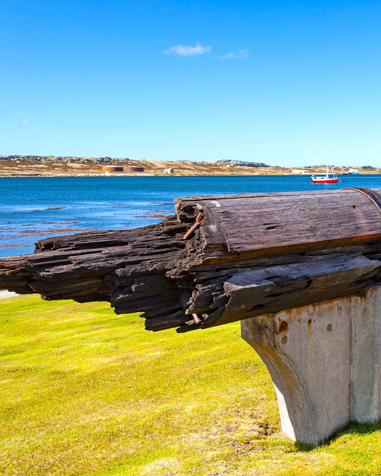 Old wooden ship mast secured with rusted metal bands, displayed on concrete supports beside a bright blue bay in the Falkland Islands.