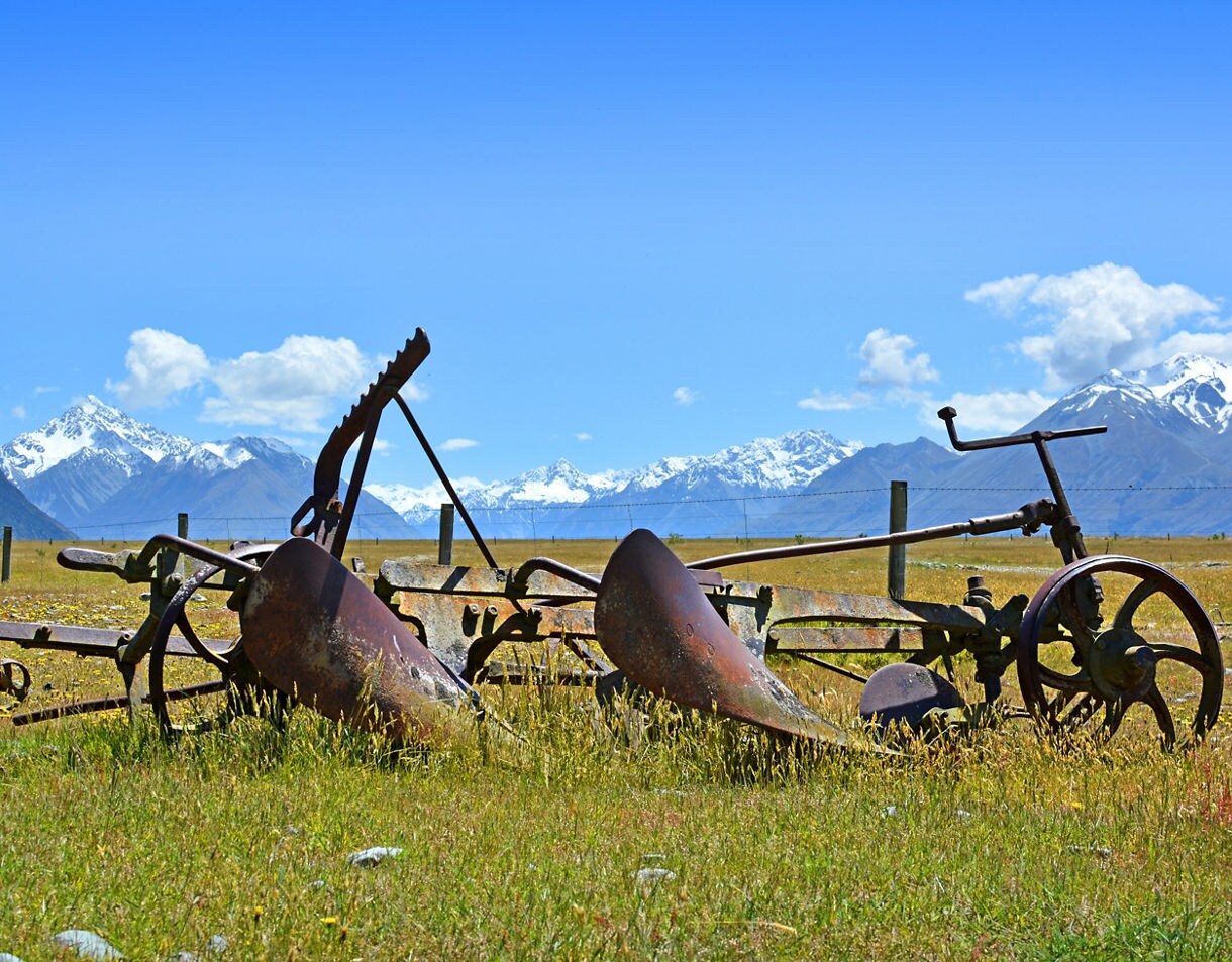 Old rusted farm equipment resting in a grassy field with tall snowy mountains rising sharply in the background under a bright blue sky.