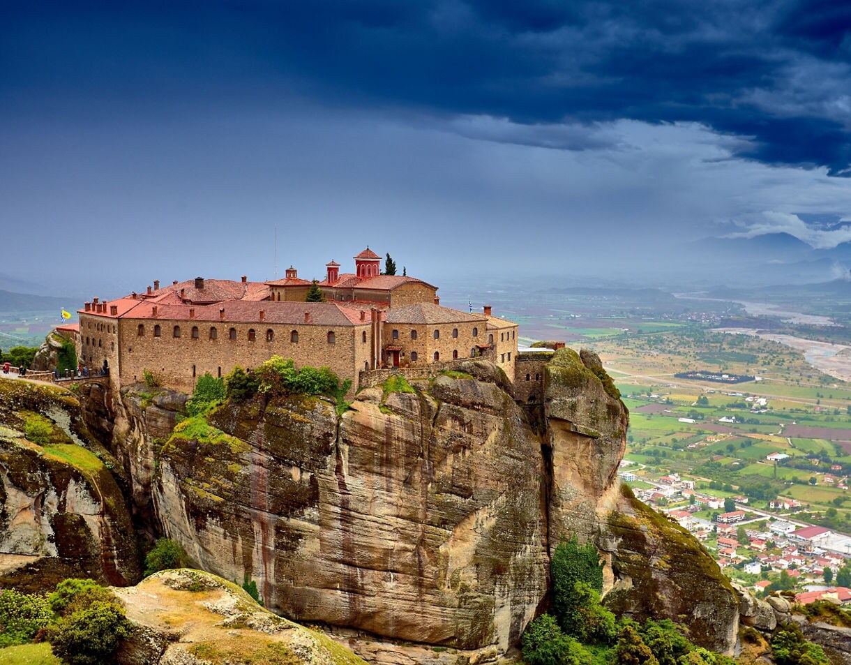 A monastery perched atop a massive rocky pillar in Meteora, Greece, overlooking a wide valley of fields and distant mountains under a dramatic sky.
