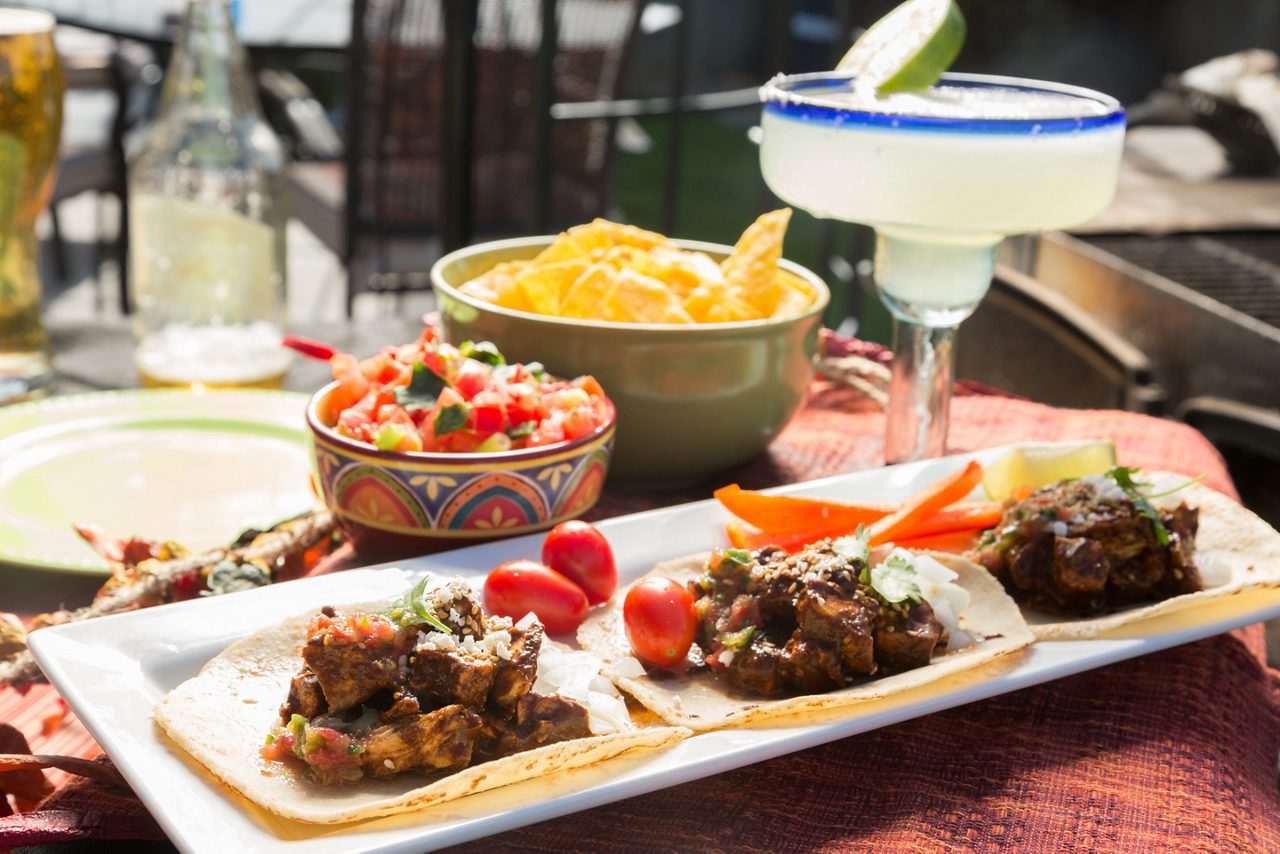 A plate of tacos filled with seasoned meat, served with cherry tomatoes, fresh salsa, tortilla chips, and a margarita garnished with a lime slice, set on an outdoor table in the sun.