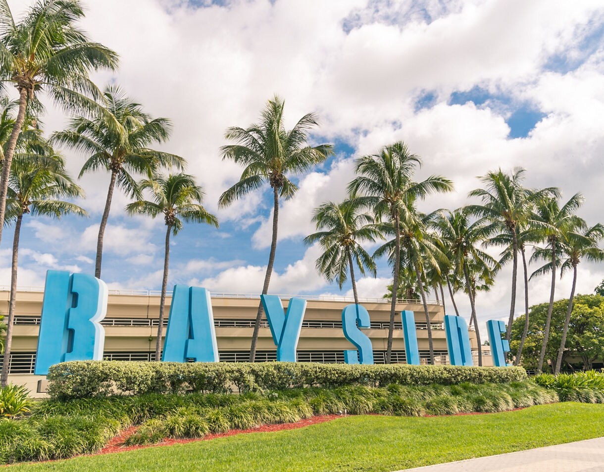 Large blue letters spelling "BAYSIDE" are set in a manicured green lawn and hedge, backed by a parking structure and several tall palm trees under a bright cloudy sky.