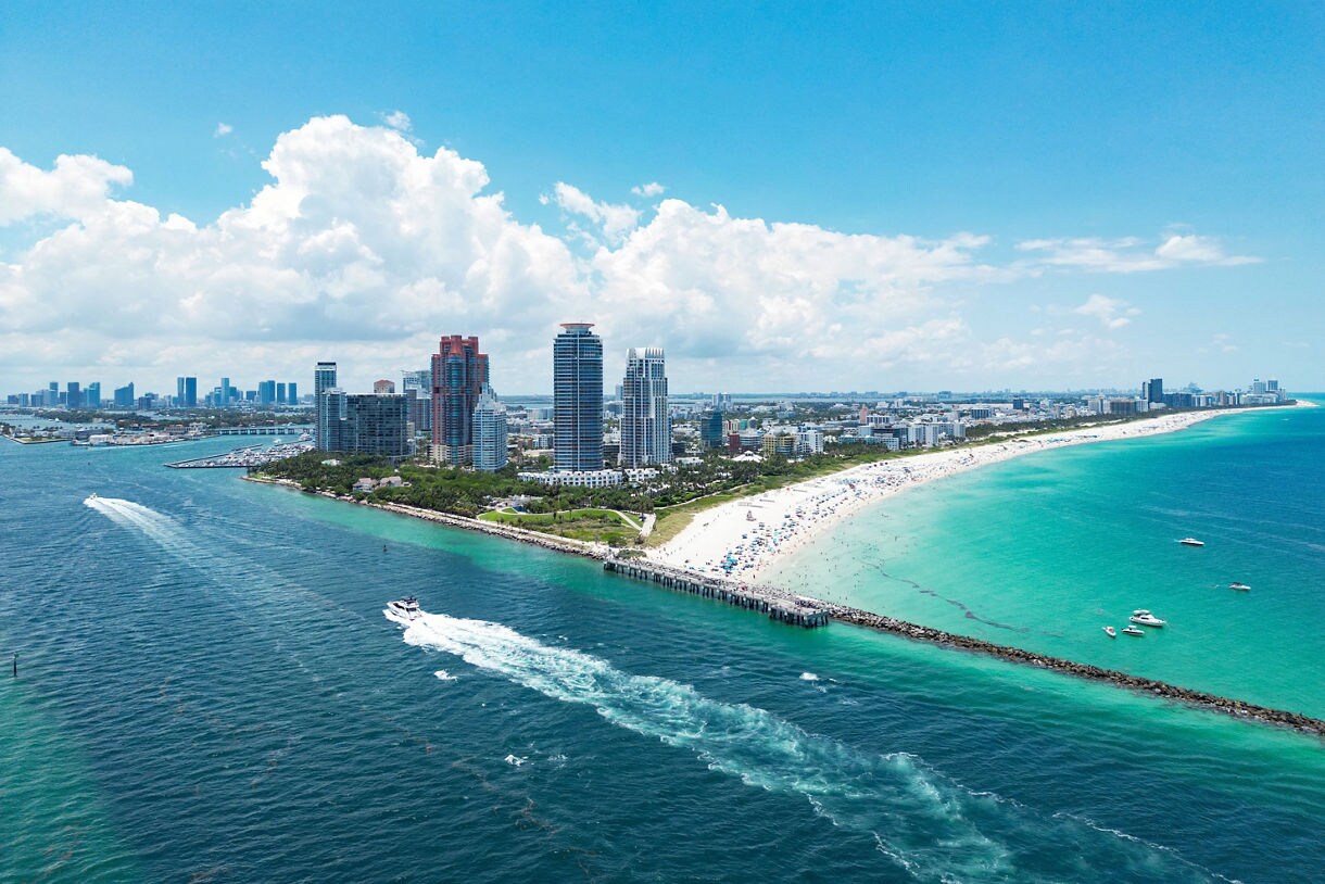 Aerial view of Miami Beach showing turquoise ocean waters, a sandy shoreline packed with beachgoers, a long pier, high-rise buildings and boats cutting through the water.