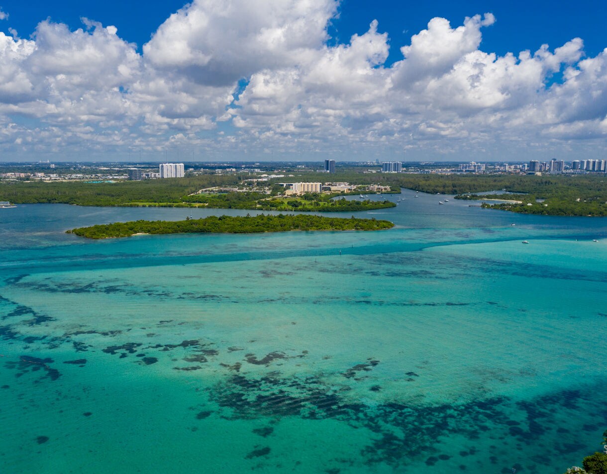 Aerial view of the stunning turquoise and teal waters of Biscayne Bay, showing a small tree-covered island in the middle ground, with the distant Miami coastline and city skyline on the horizon under a bright cloudy sky.