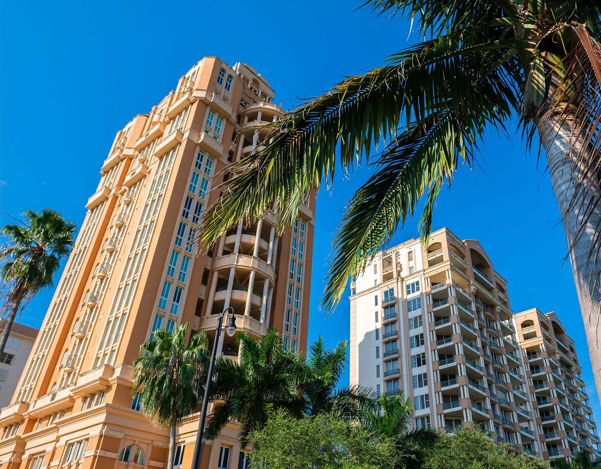 Low-angle view of two tall, peach-colored Mediterranean Revival style apartment buildings in Coral Gables Florida, framed by palm trees against a brilliant blue sky.