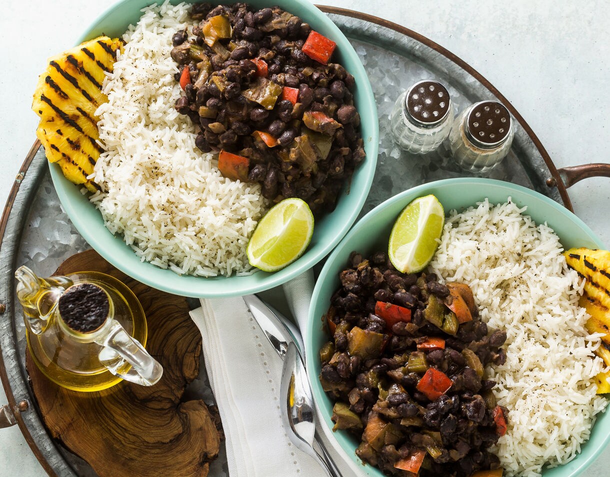 Overhead view of two teal bowls containing white rice and Cuban-style black beans mixed with red and green bell peppers, each served with a lime wedge and a grilled pineapple slice on a rustic metal tray.
