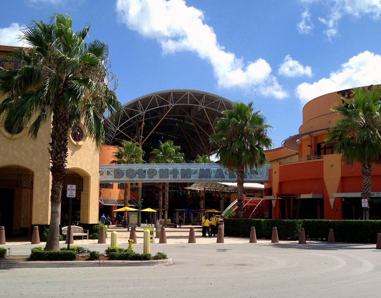 The outdoor entrance to Dolphin Mall, featuring orange-colored buildings, palm trees, and a large arched metal and fabric canopy over the walkway beneath a blue sky with white clouds.