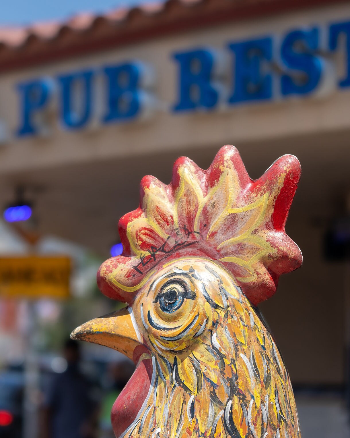 Close-up of a colorful yellow and red painted ceramic rooster statue with a blue eye, with the blurred sign of "El Pub Restaurant" visible in the background in Little Havana.