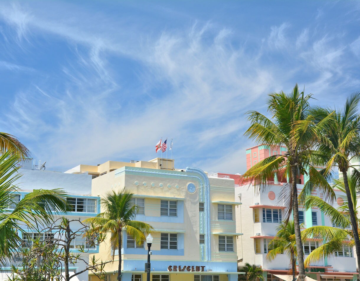 A low-angle view of colorful Art Deco hotels on Miami's South Beach, with palm trees framing the buildings against a bright blue sky with wispy white clouds.