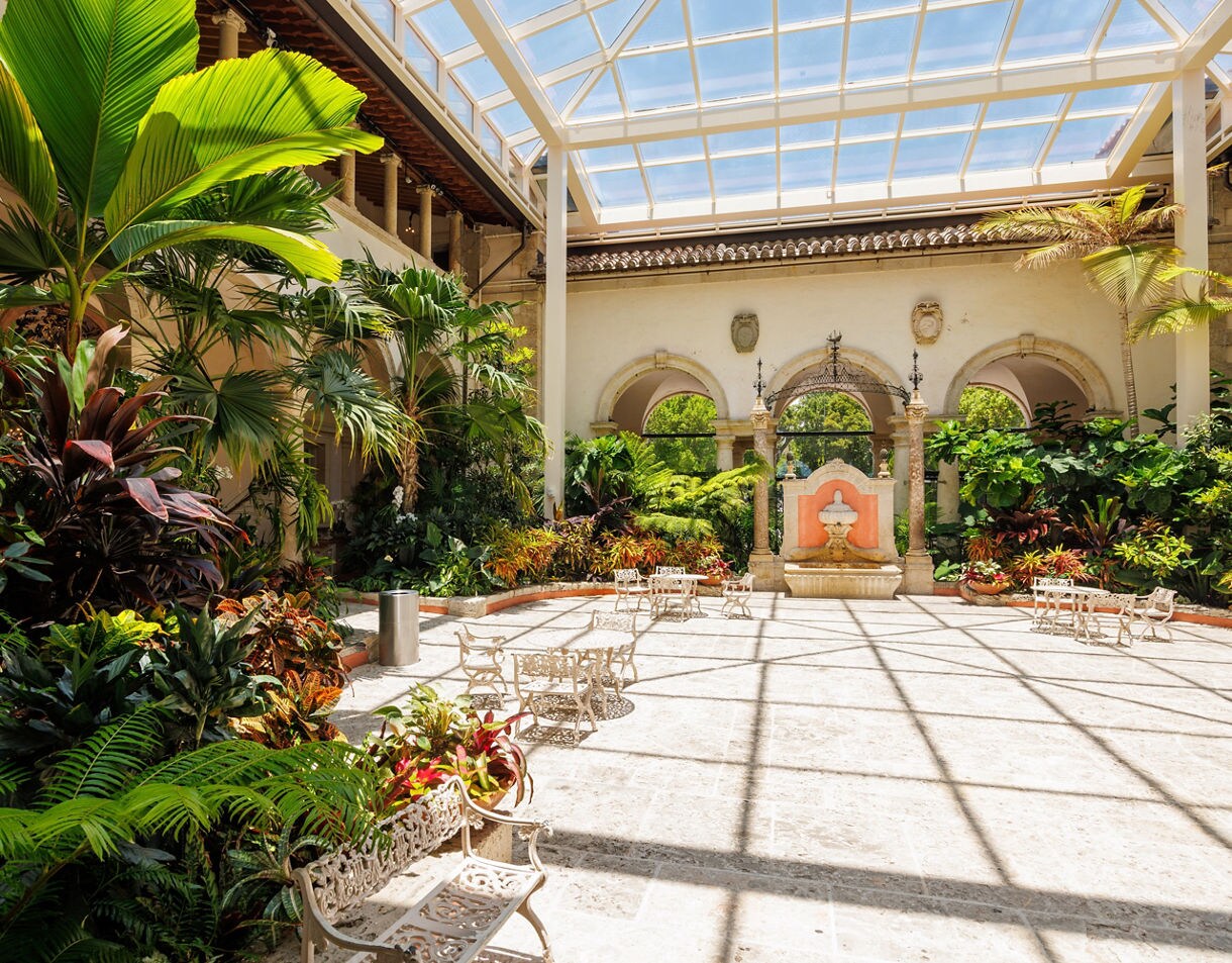 The enclosed courtyard of the Vizcaya Museum and Gardens features a glass roof, tropical plants, a small fountain, and white wrought iron tables and chairs on the stone floor.