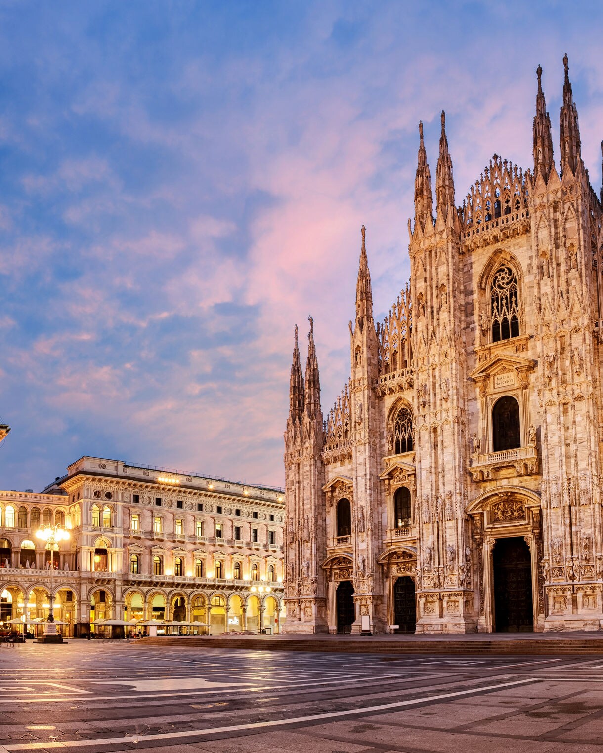 Wide view of Milan’s Duomo at dusk, showing its ornate gothic spires and marble exterior glowing in warm evening light, with the surrounding square and gallery arches nearby.