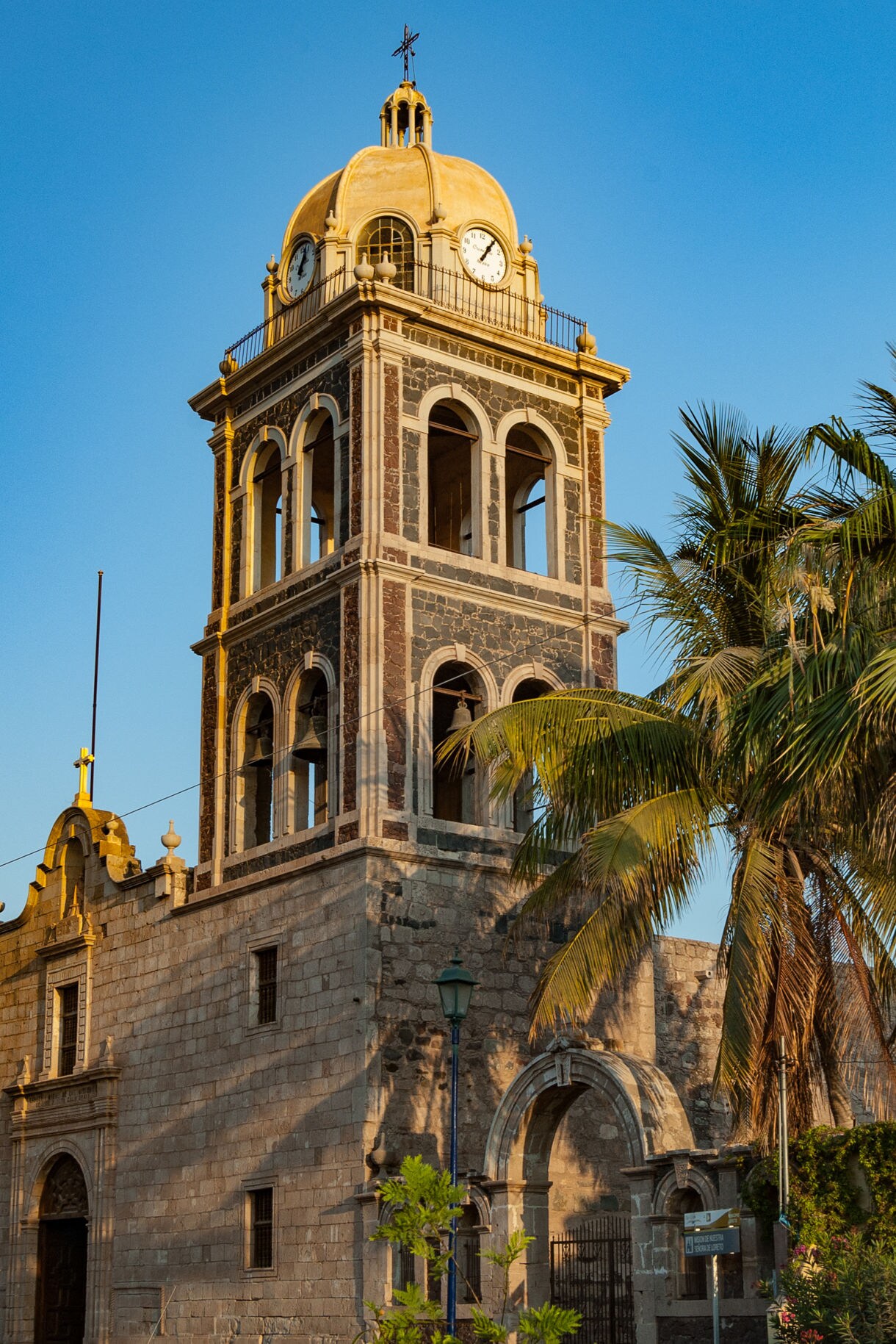 Historic stone church in Loreto with a tall clock tower and golden dome, framed by palm trees under a clear sky.