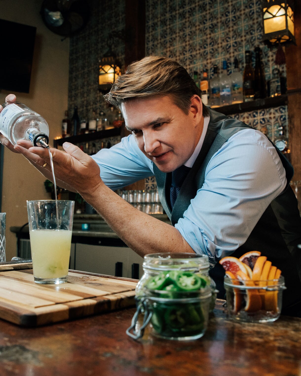 Bartender Rob Floyd in vest and shirt pouring a cocktail with fresh garnishes and ingredients displayed on bar. 