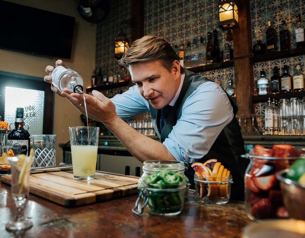 Bartender Rob Floyd in vest and shirt pouring a cocktail with fresh garnishes and ingredients displayed on bar.