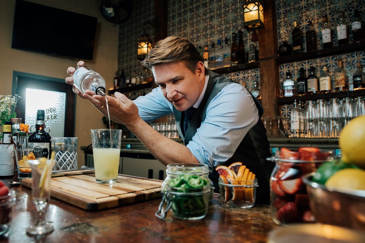 Bartender Rob Floyd in vest and shirt pouring a cocktail with fresh garnishes and ingredients displayed on bar. 