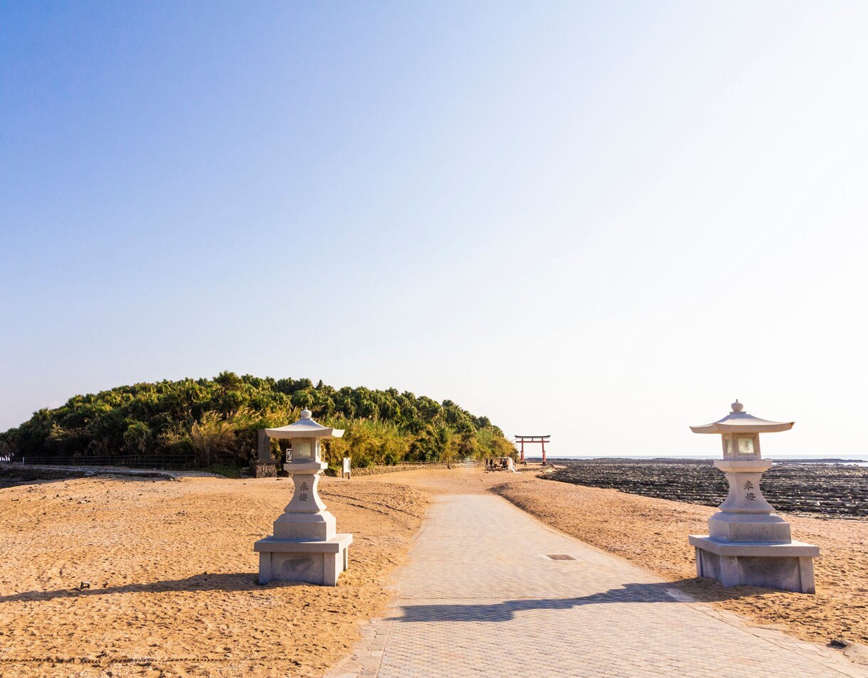 Stone lanterns flanking a sandy path leading to Aoshima Island and a red torii gate by the ocean in Miyazaki, Japan.