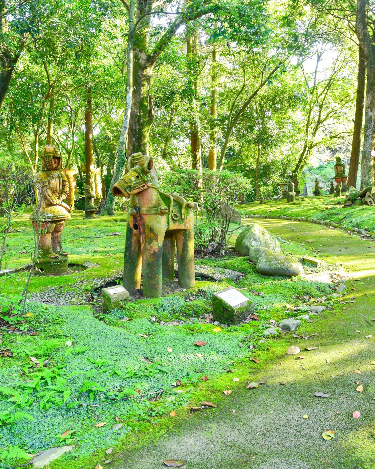 Haniwa Garden in Miyazaki, Japan, featuring clay warrior and horse statues scattered throughout a mossy forest landscape with a winding shaded path.