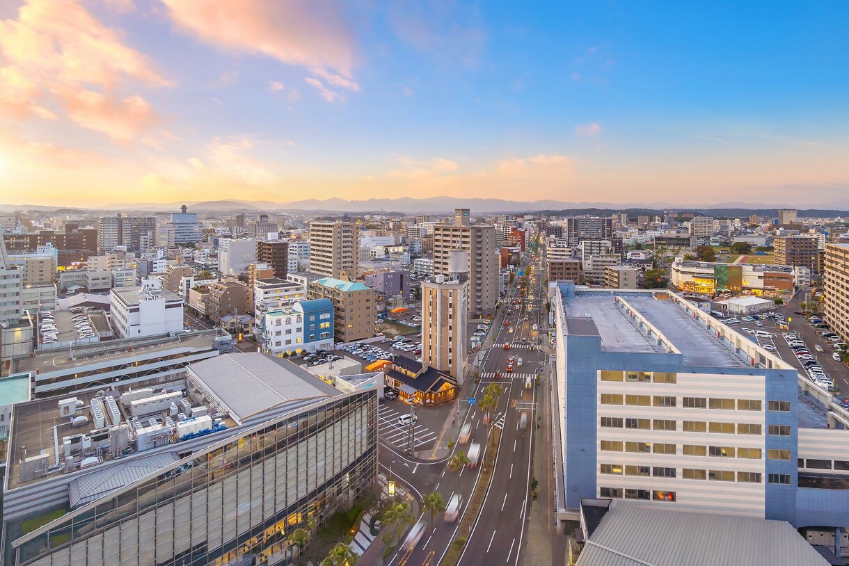 Panoramic view of Miyazaki, Japan at sunset with city buildings, roads, and soft golden light over the skyline.