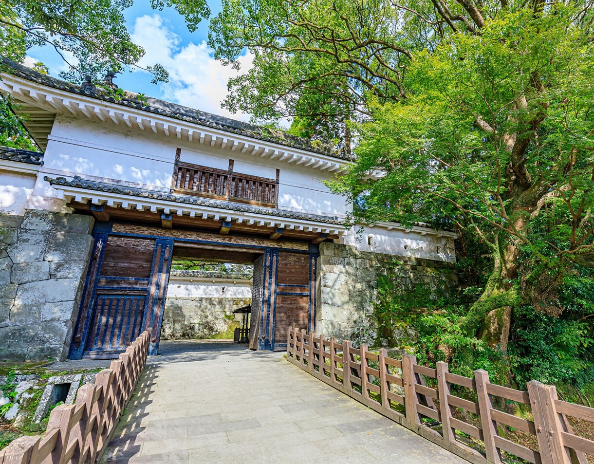 Entrance gate of Obi Castle in Miyazaki, Japan, featuring white walls, wooden doors, stone foundations, and lush green trees surrounding the pathway.
