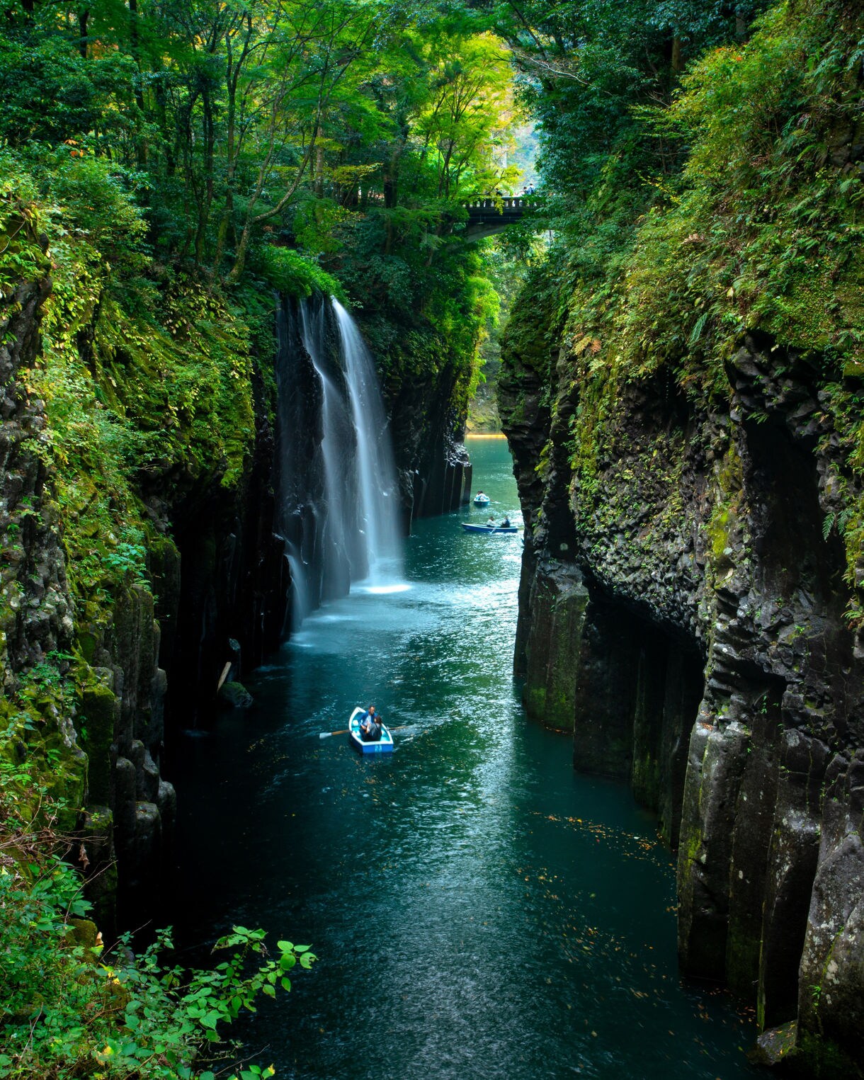 A tranquil river cutting through high rocky cliffs with a waterfall and boats traveling beneath forested surroundings.