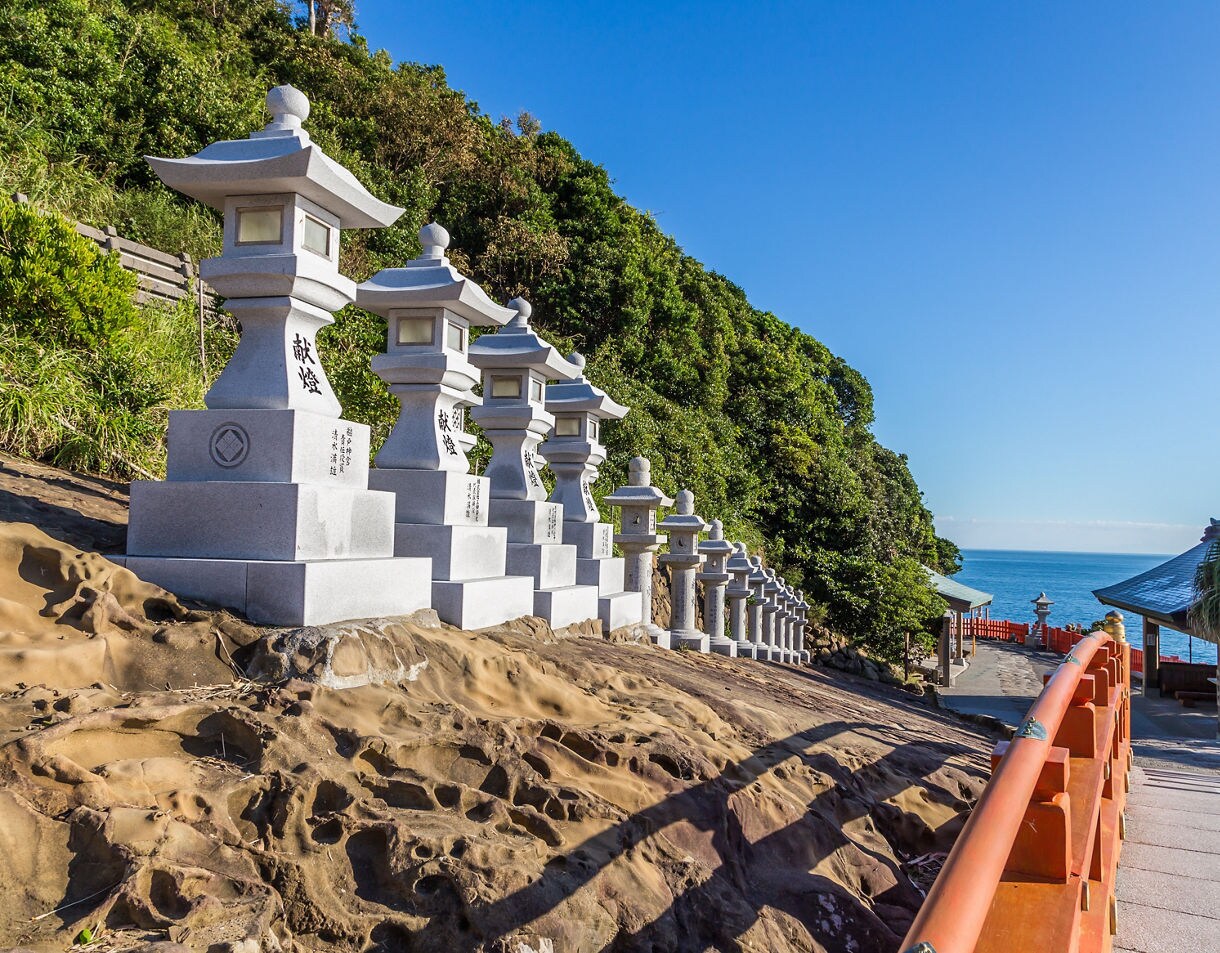 Stone lanterns lined up along a coastal shrine pathway in Miyazaki, Japan, with ocean views and red railings.