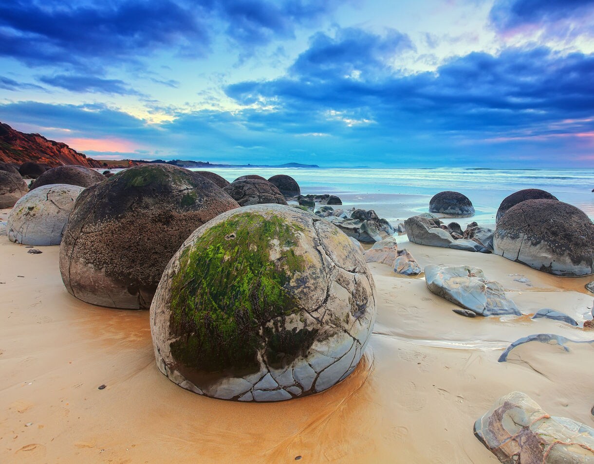 Spherical Moeraki Boulders scattered across a sandy beach at sunrise, with patches of green moss and gentle waves rolling in under a colorful sky.