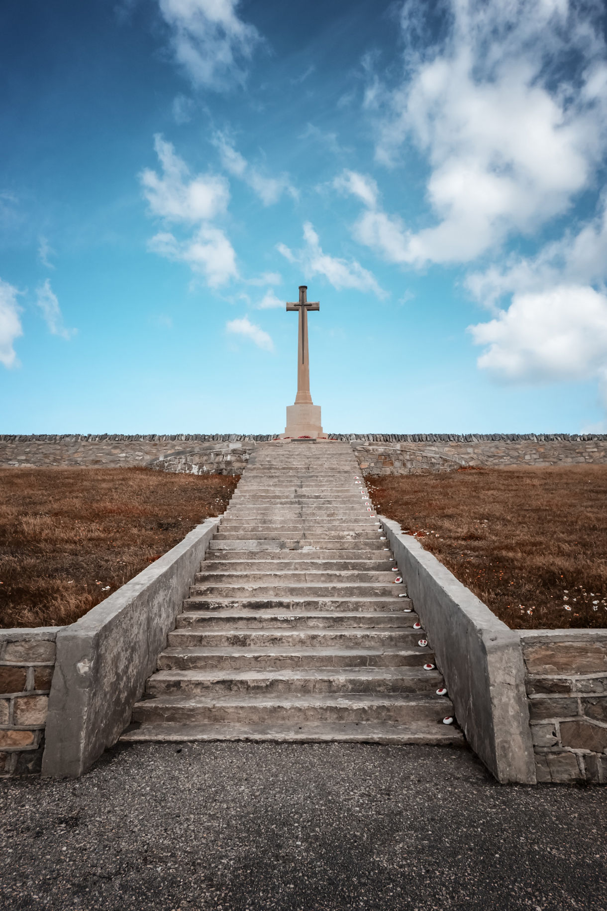 Stone staircase leading up to a tall memorial cross on a hilltop, framed by a bright blue sky with scattered clouds.