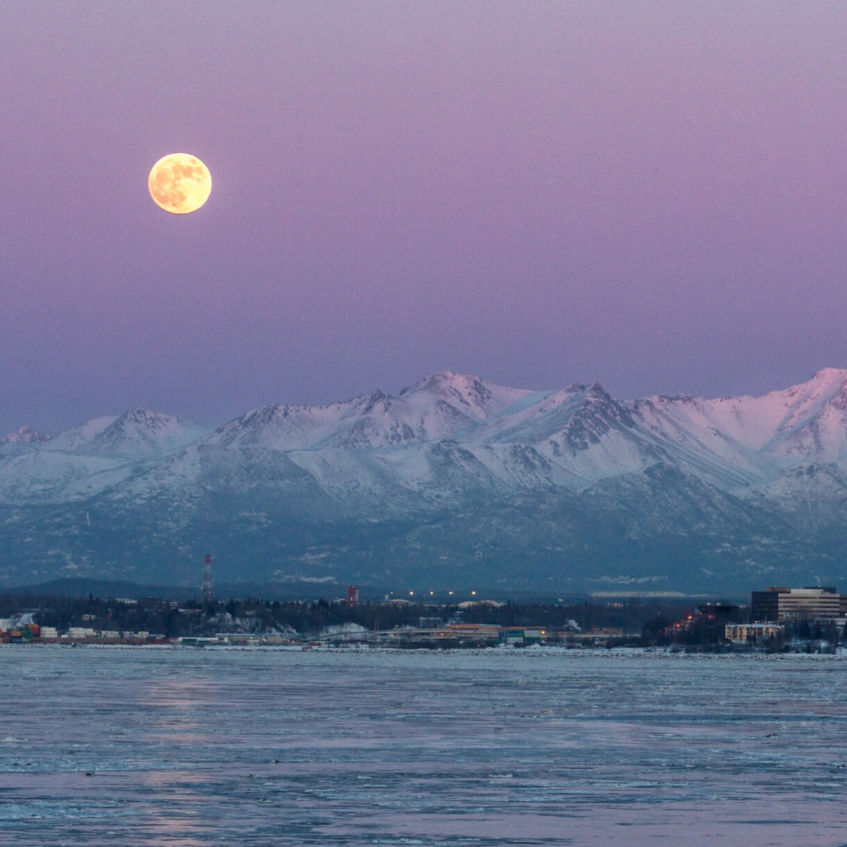 Full moon over Anchorage, Alaska skyline with snow-covered Chugach Mountains in the background during twilight.