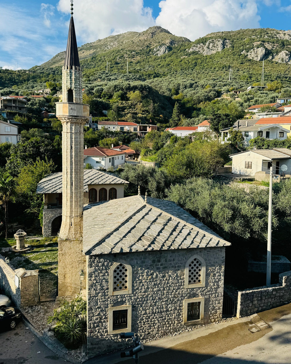 Stone mosque with a tall slender minaret set among olive trees and red-roofed homes, backed by green mountains under a bright blue sky.