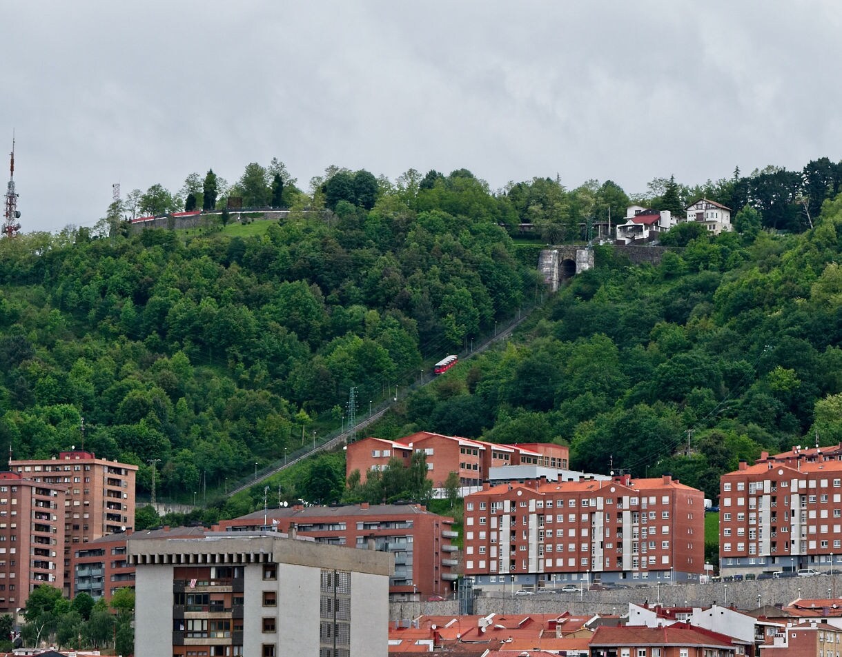 View of Bilbao’s hillside with red-roofed apartment buildings and a funicular railway ascending through dense green forest toward Mount Artxanda.